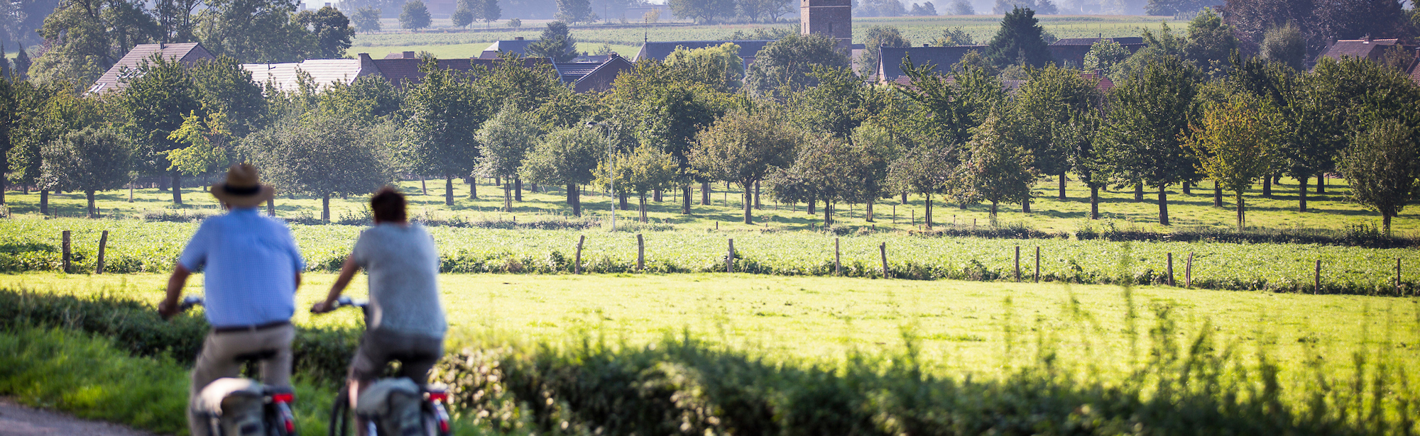 Koppel fietst langs weiland met uitzicht op de kerk en het dorpje Mesch